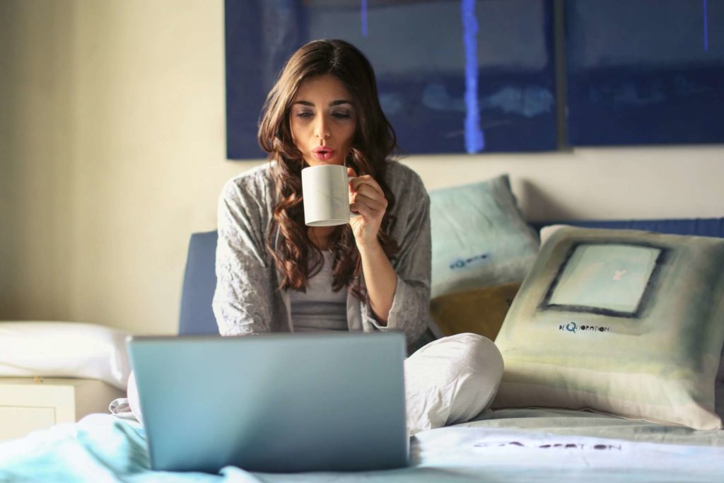 A young woman sitting on her bed, blowing on her hot coffee, and looking at her laptop