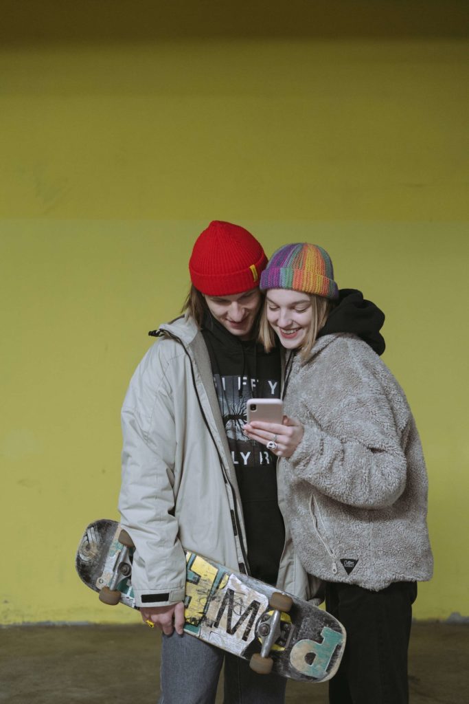 Two teenagers wearing beanies smiling and looking at the girl's phone