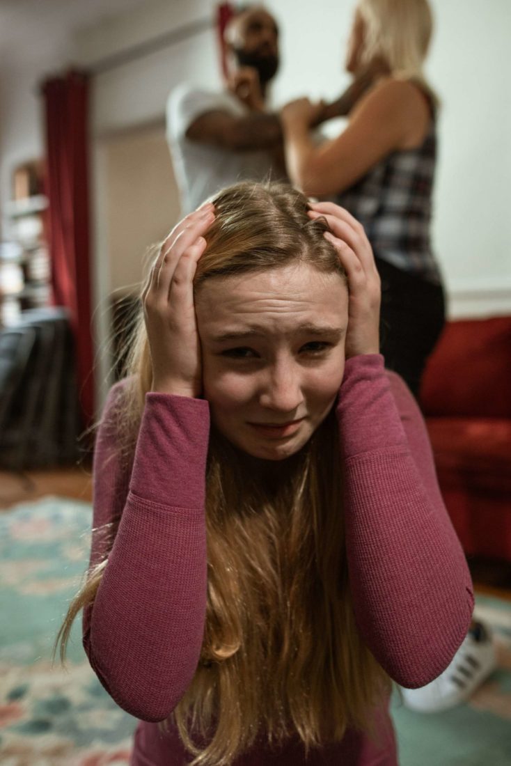 A young girl crying and covering her ears while her parents fight in the background