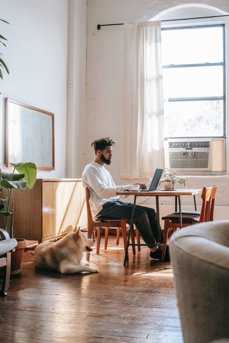 A young black man with his dog sitting at a table on his laptop