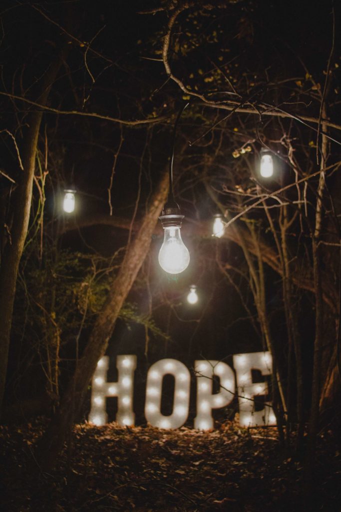 Lit-up letters that spell "Hope" outside in a forest with light bulbs handing on nearby branches