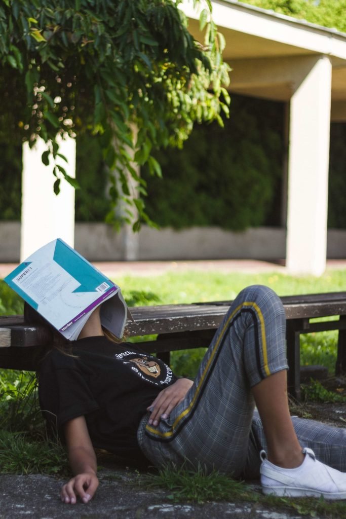 A young woman laying against a bench with a textbook covering her face