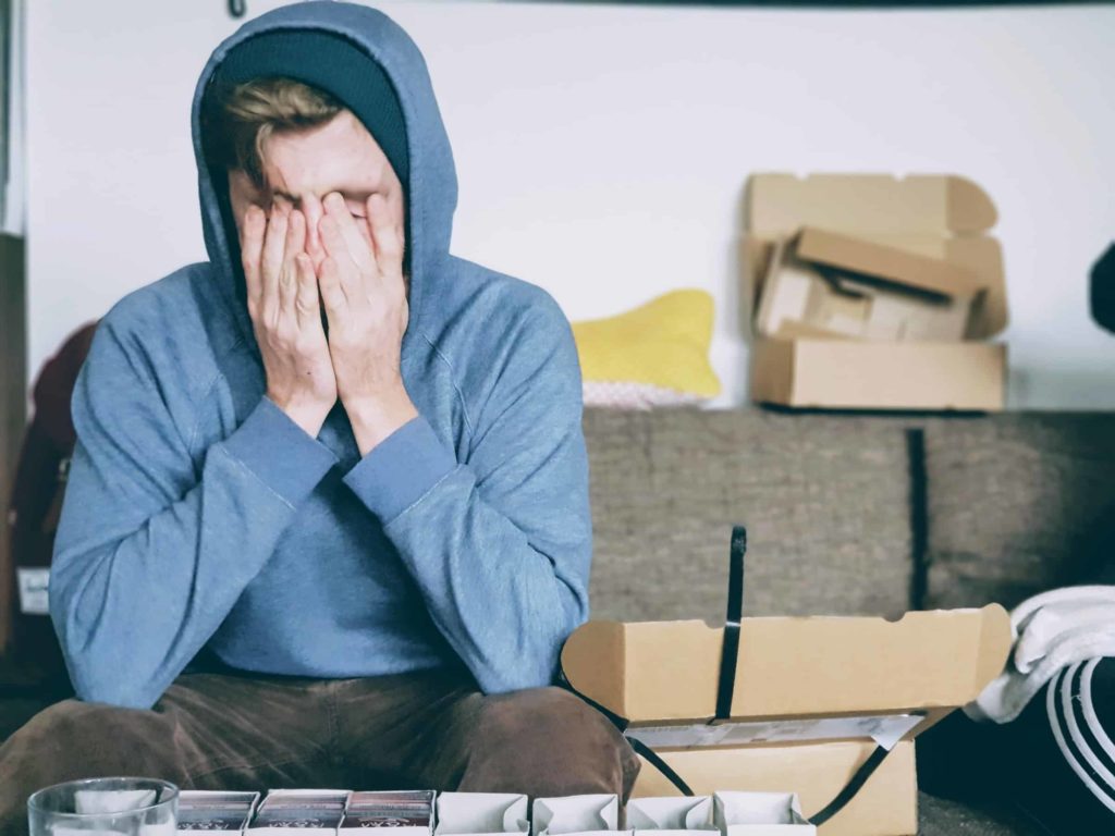 A stressed young white man with his hands on his face surrounded by moving boxes