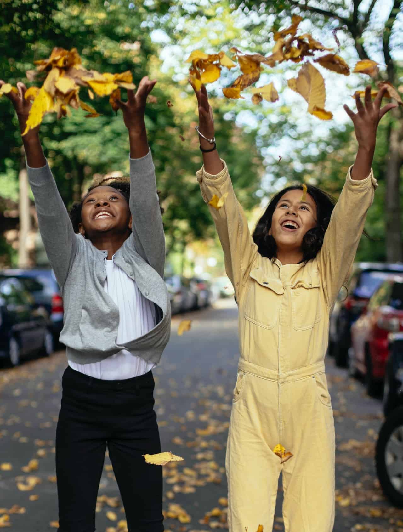 A young black girl and young Hispanic girl throwing leaves in the area in the street