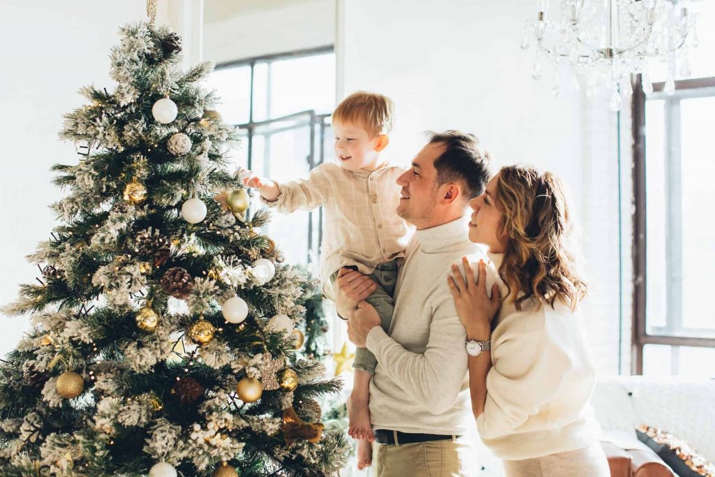 A mom and dad holding their child while he puts an ornament on the Christmas tree