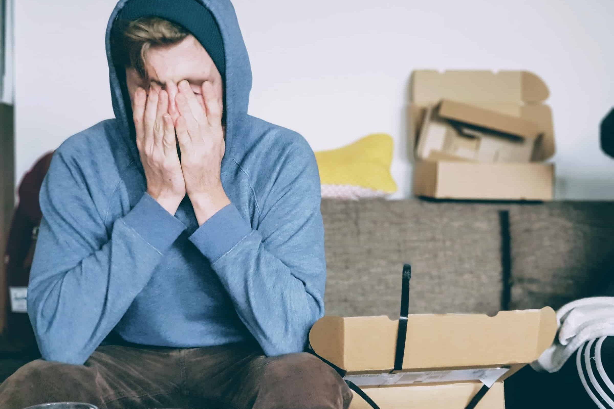 A stressed young white man with his hands on his face surrounded by moving boxes